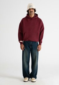 Uomo che indossa un berretto lavorato a maglia color crema, una felpa borgogna, jeans larghi blu scuro e sneaker color crema, in piedi davanti a uno sfondo bianco semplice.