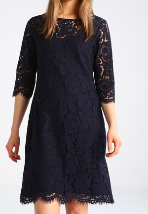 Kobieta ubrana w granatową koronkową sukienkę sięgającą do kolan, z rękawami do połowy przedramienia i kwiatowym wzorem koronki.