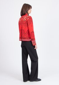 Röd broderad jacka med svarta mönsterdetaljer, kombinerad med svarta vida byxor. Jackan har långa ärmar och hög krage.