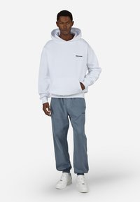 Λευκή κουκούλα με μπροστινή τσέπη και μαύρο λογότυπο, συνδυασμένη με ανοιχτό μπλε παντελόνι με κορδόνια και λευκά αθλητικά παπούτσια. Απλός σχεδιασμός, χαλαρή εφαρμογή.