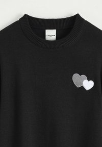 Pull en tricot noir avec un col rond, comportant deux cœurs brodés en gris et blanc sur la partie gauche de la poitrine.