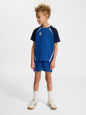 Barn med krøllet hår iført en blå sportstrøje og shorts, hvide sokker og hvide sneakers, står og kigger til siden.