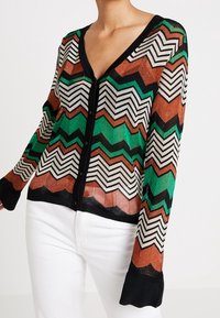 Kvinna iklädd en knäppt kofta med zigzagmönster i svart, grönt, vitt och rostfärgat, kombinerat med vita byxor.