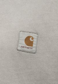 Ανοιχτό γκρι πλεκτό ύφασμα με ένα μικρό τετράγωνο λογότυπο Carhartt ραμμένο στο κέντρο σε καφέ και λευκές αποχρώσεις.