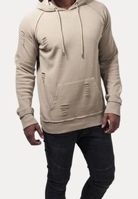 Beige distressed hoodie with a kangaroo pocket, drawstring hood, and raglan sleeves. Features frayed details and ribbed cuffs. Worn with black jeans.