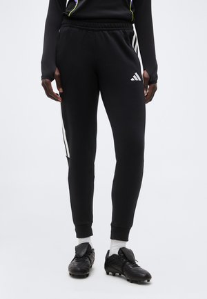 Άτομο που φοράει μαύρο παντελόνι φόρμας Adidas με άσπρες ρίγες και λογότυπο, άσπρες κάλτσες και μαύρα παπούτσια ποδοσφαίρου, σε απλό φόντο.