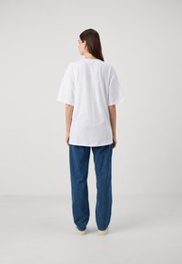 White cotton t-shirt with short sleeves and a relaxed fit, paired with blue denim pants. Simple, unadorned design, and slightly longer back.