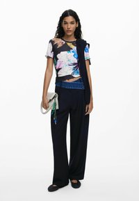 Top corto de manga corta con estampado floral sobre una base negra, combinado con pantalones anchos oscuros. Lleva un bolso gris con acentos verdes y herrajes azules.