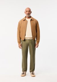 Bruine bomberjack, crèmekleurige gebreide trui, olijfgroene broek en bruine sneakers met zwarte accenten. Lacoste-logo op de jack.