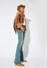 Tan suede fringed jacket, valkoinen paita, vaaleansiniset flare-farkut revityillä helmoilla ja tan cowboy-kengät. Hattu ja asusteet viimeistelevät ilmeen.