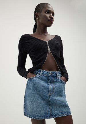 Schwarze, gerippte Zip-Up-Strickjacke mit langen Ärmeln, kombiniert mit einem hellblauen Jeans-Minikleid mit Knopfverschluss und Seitentaschen.