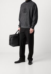 Sweat-shirt en maille gris foncé avec logo, pantalon noir, et chaussures noires polies. Porte un sac de voyage en cuir noir texturé.