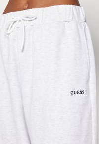 Svijetlosivi dukserici hlače od mekanog materijala, s elastičnom gumicom i vezicom. Crni "GUESS" logo na lijevoj nozi.