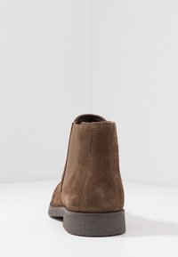Botín de ante marrón hasta el tobillo con paneles laterales elásticos y suela de goma texturizada. El diseño es sencillo, con líneas limpias y costuras mínimas.