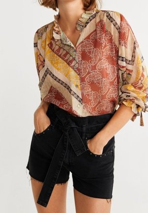 Femme portant un chemisier à manches longues à motifs rentré dans un short en jean noir taille haute avec une ceinture nouée et les mains dans les poches.