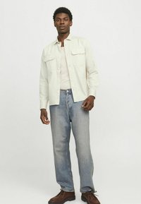 Camicia chiara con bottoni e due tasche sul petto, abbinata a jeans azzurri e scarpe stringate marroni. Design semplice e informale.