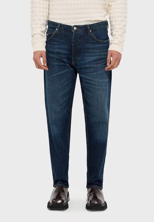 Jeans in denim blu scuro con un design a gamba dritta, cinque tasche e cuciture in rame. Abbinati a scarpe in pelle nere e a un maglione in crema con texture.