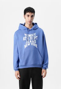Blue hoodie made of cotton with a front kangaroo pocket. Features white text “TOMMY ATH. DEPT. JEANS NEW YORK” across the chest.