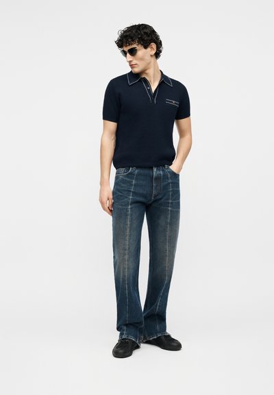 Bărbat purtând un polo negru cu mâneci scurte tricotat, blugi largi decolorati, pantofi negri și ochelari de soare, stând cu o mână în buzunar pe un fundal alb.