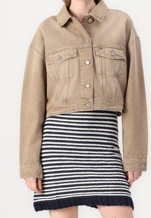Beige gekürzte Jeansjacke mit Metallknöpfen, kombiniert mit einem schwarz-weiß horizontal gestreiften Strickrock mit Rüschensaum.