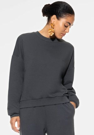 Dunkelgrauer Sweatshirt mit langen Ärmeln, rundem Halsausschnitt und lockerer Passform. Das Material hat eine glatte Textur. Kombiniert mit großen, dekorativen Ohrringen.