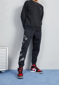 Sudadera y pantalones deportivos negros con logo blanco, combinados con zapatillas negras y rojas. Textura suave, ajuste informal y colores contrastantes.