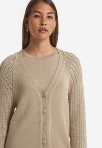 Beige stickad cardigan med V-hals, knäppning framtill och texturerade ärmar med ett öppet stickat mönster.
