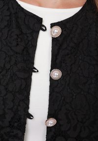 Jachetă din dantelă neagră cu modele florale, nasturi rotunzi din perle cu accente argintii și o margine neagră de-a lungul decolteului. Purtată peste un top alb.