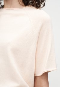 Ženska, oblečena v svetlo bež kratkotrajni pleten zgornji del z okroglim izrezom, prikazuje zgornji del trupa in del kodrastih temnih las.