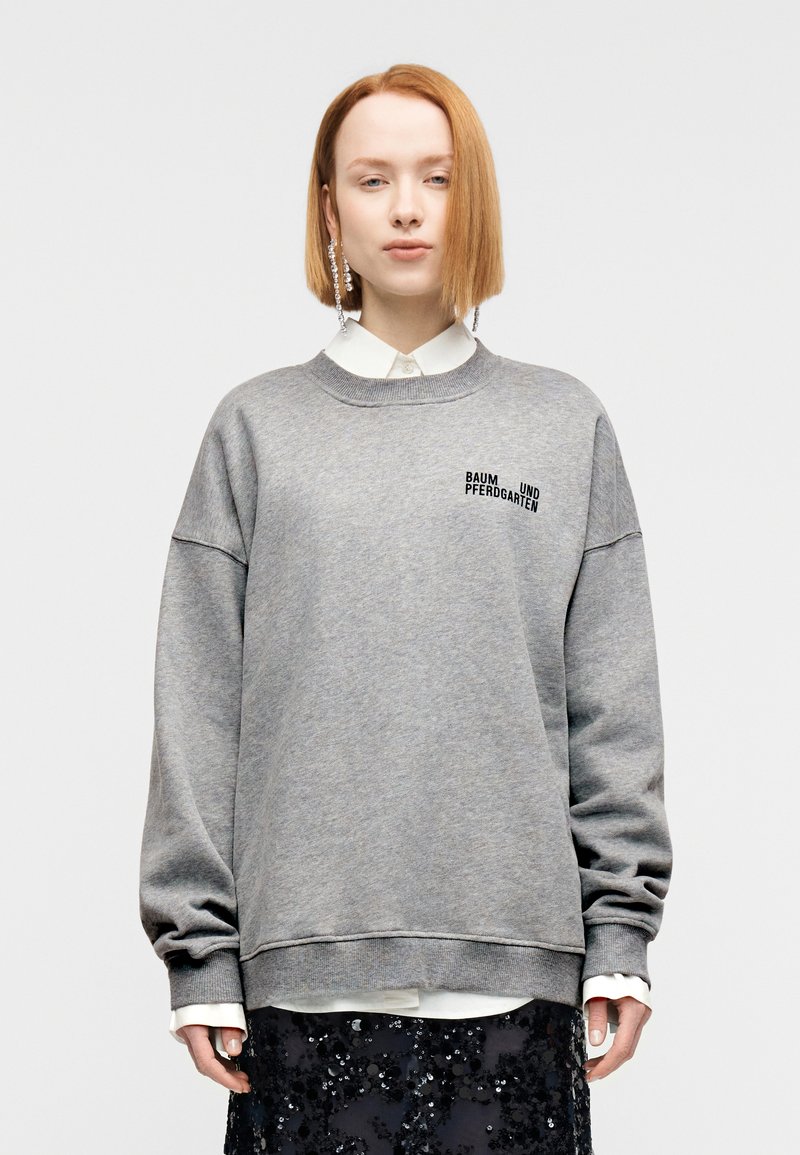 Γκρι φούτερ με στρογγυλή λαιμόκοψη, χαλαρούς ώμο υς και μαύρο τυπωμένο κείμενο. Φορεμένο πάνω από ένα λευκό πουκάμισο με γιακά, σε συνδυασμό με μια λαμπερή μαύρη φούστα με παγιέτες.