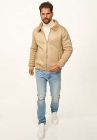 Beige Kunstfelljacke mit Reißverschluss, gerippten Bündchen und warmem Kragen, kombiniert mit hellblauen Jeans und weißen Turnschuhen.