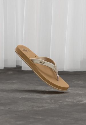 Jasnobrązowa i beżowa klapka na gumce z teksturowaną podeszwą i materiałowymi paskami, umieszczona na szarym tle podłogi i białej zasłony.