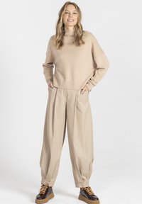 Beżowy sweter dzianinowy z wysokim kołnierzem, w zestawieniu z luźnymi beżowymi spodniami i ciemnobrązowymi trampkami z teksturowanymi akcentami.