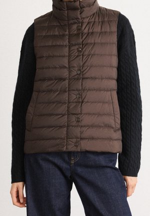 Personne portant un gilet matelassé marron boutonné par-dessus un pull noir à torsades et un jean bleu foncé, debout devant un fond neutre.