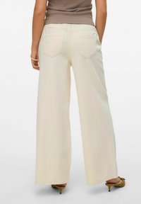 Plati-beigās bikses ar gludu tekstūru, elastīgu jostu un divām aizmugures kabatām ar rombveida atloka dizainu. Minimālas šūšanas detaļas.