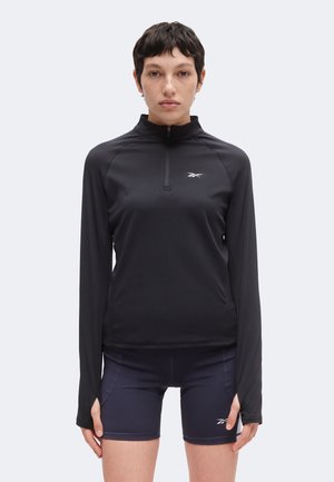 Osoba z krótkimi ciemnymi włosami ma na sobie czarną bluzę na zamek z długim rękawem i granatowe sportowe szorty, oba elementy z małymi białymi logo Reebok, stoi przodem.