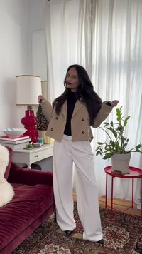 Γυναίκα με μακριά σκουρόχρωμα μαλλιά, ντυμένη με μπεζ σακάκι, μαύρο τοπ και λευκό παντελόνι, στέκεται σε φωτεινό δωμάτιο με κόκκινο βελούδινο καναπέ, φυτό και λευκές κουρτίνες.