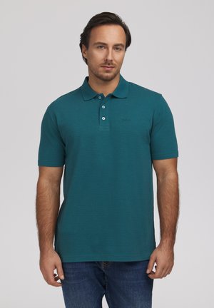 Mężczyzna z krótkimi ciemnymi włosami, ubrany w turkusową koszulkę polo z krótkim rękawem i niebieskie dżinsy, stoi na tle jednolitego jasnego tła.