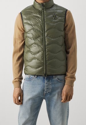 Homme portant une veste matelassée vert olive sur des manches longues beige et un jean bleu clair, debout devant un fond uni.