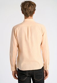 Langärmeliges Hemd aus strukturiertem, hellpfirsichfarbenem Stoff, mit Kentkragen, einem einfachen Rückenfalten und geknöpften Manschetten. Sichtbare schwarze Jeans.