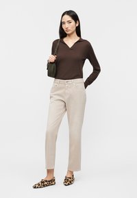 Femme debout portant un haut marron à manches longues, un pantalon beige, des chaussures à imprimé léopard, et portant un petit sac à bandoulière vert foncé.