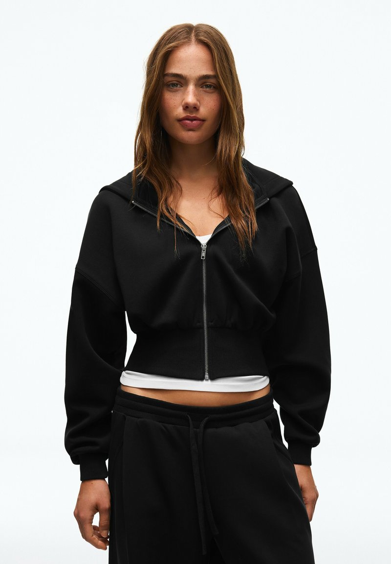 Jeune femme aux longs cheveux bruns portant un sweat à capuche court noir zippé et un pantalon noir, debout devant un fond clair uni.
