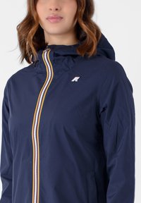 Giacca navy con cappuccio, caratterizzata da una cerniera frontale evidenziata da righe multicolori e un piccolo logo sul petto. Texture liscia.