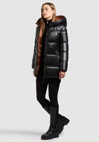 Pufferjack met een zwarte buitenkant en een bruine binnenvoering, voorzien van een capuchon, een ritssluiting aan de voorkant en een gestikte uitstraling. Gekleed met zwarte leggings en laarzen.