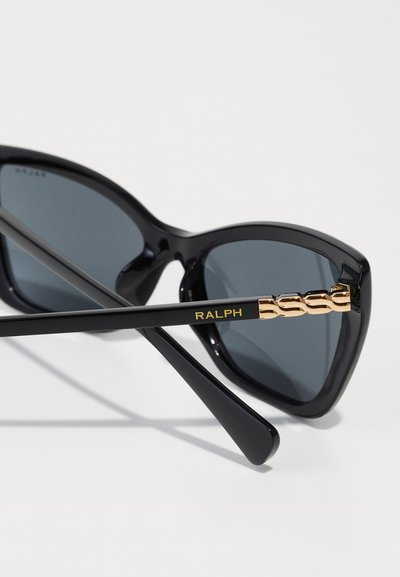 Gafas de sol negras con forma de ojo de gato, lentes oscuros y acentos dorados en las monturas. El brazo presenta "RALPH" en letras doradas.