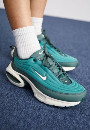 Stopy w turkusowo-ciemnozielonych sneakersach Nike z białymi podeszwami i białych, prążkowanych skarpetkach na niebieskim dywanowym podłożu.
