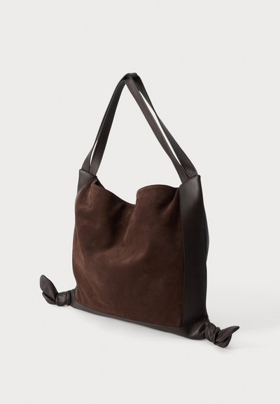 Sac fourre-tout en daim marron foncé avec des poignées en cuir lisse et des détails en cuir noués aux coins inférieurs sur un fond clair.