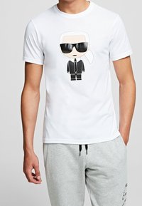 Camiseta de manga corta blanca con un personaje de dibujo animado con coleta, traje negro y gafas de sol en un estampado gráfico. Tejido de algodón.