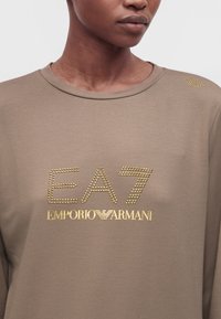 Femme portant un sweat-shirt marron avec des clous dorés formant EA7 et un logo Emporio Armani imprimé sur la poitrine et l'épaule.