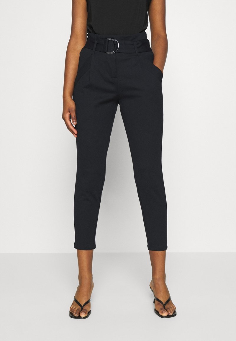 Femme portant un pantalon noir court taille haute avec ceinture et sandales noires à talons, debout avec une main dans la poche sur fond blanc.